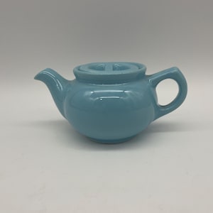 May include: A small, light blue ceramic teapot with a matching lid. The teapot has a rounded body, a curved spout, and a loop handle. The teapot is a solid color and has a glossy finish. The teapot is designed for brewing tea.
