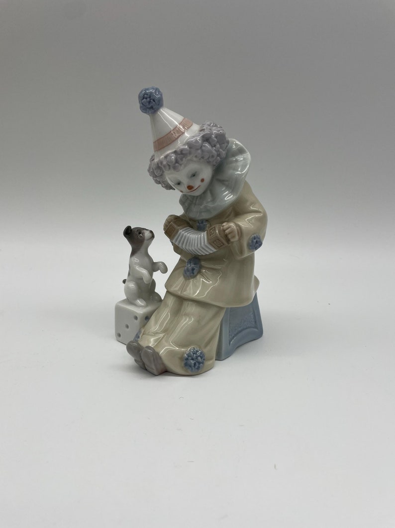 May include: A porcelain figurine of a clown playing an accordion with a small dog sitting on a dice block. The clown is wearing a colourful costume with a blue and white hat.