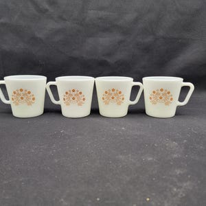 May include: Four white ceramic mugs with brown floral designs. The mugs have a rounded shape and a handle on the side.