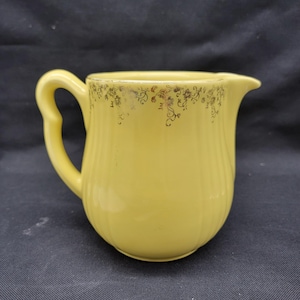 May include: A yellow ceramic pitcher with a gold floral design around the top rim. The pitcher has a rounded body and a handle.