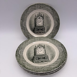 May include: A stack of three round ceramic plates featuring a dark green transferware pattern. Each plate displays a central illustration of a grandfather clock with Roman numerals and a decorative border with a cross-hatched texture.