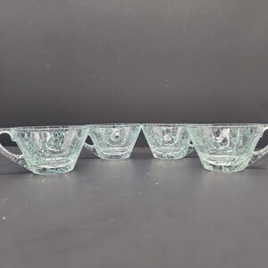 May include: Set of four clear glass teacups with a crackle glaze finish. The cups have a rounded shape and a handle on one side.
