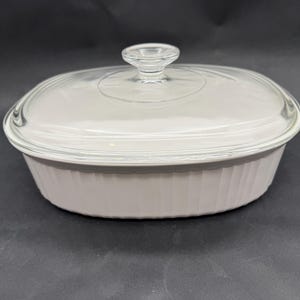 May include: An oval white ceramic baking dish with vertical ribbed sides and a clear glass lid featuring a central knob handle. The dish has a smooth, opaque finish and the lid is transparent, allowing a view of the interior rim of the vessel.