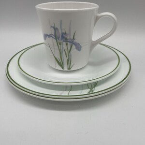 May include: A white ceramic mug with a blue and green floral design, resting on two white plates with green trim. The mug has a handle and a slightly flared shape. The plates are stacked, with the top plate smaller than the bottom one.