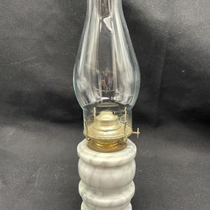 May include: An antique oil lamp with a clear glass chimney and a brass burner. The base is made of white marble with gray veining. The lamp is designed to sit on a flat surface.