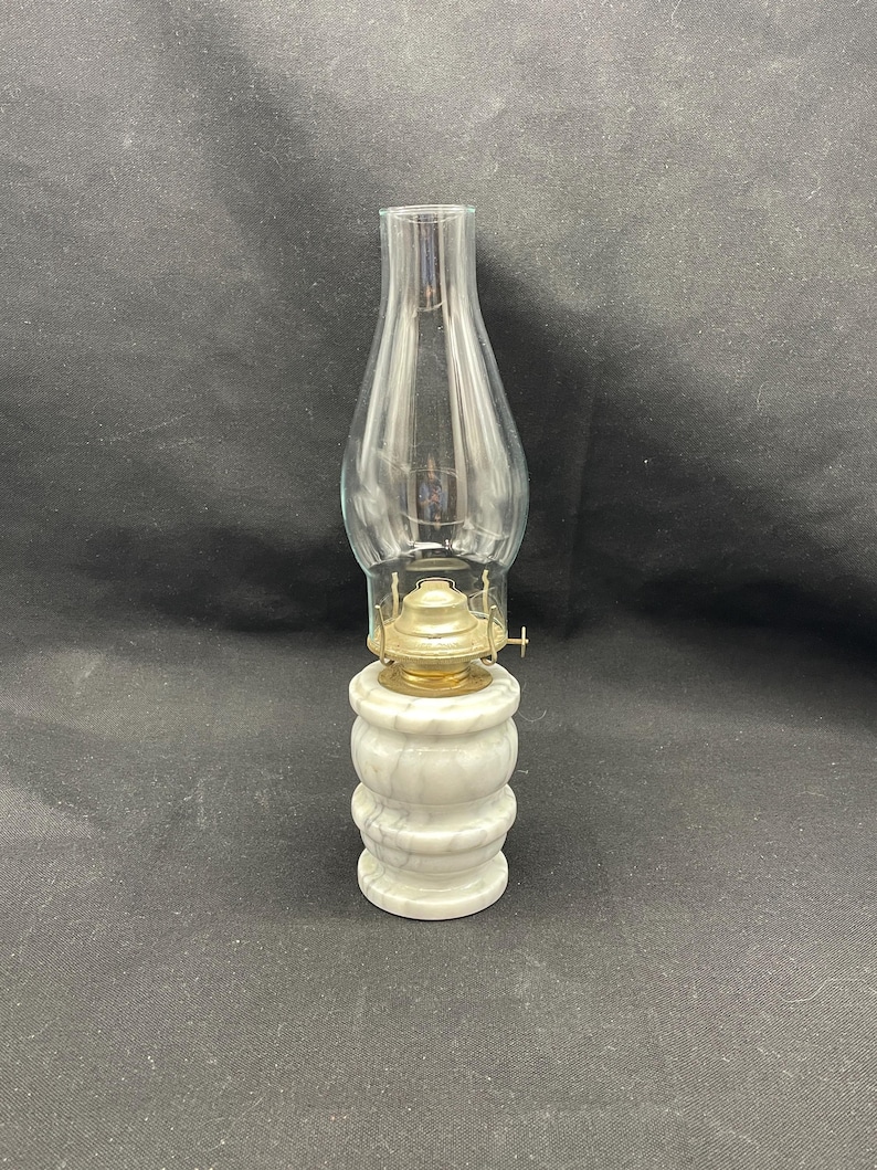 May include: A vintage oil lamp with a clear glass chimney and a brass burner. The base is made of white marble with gray veining. The lamp is designed to sit on a flat surface.