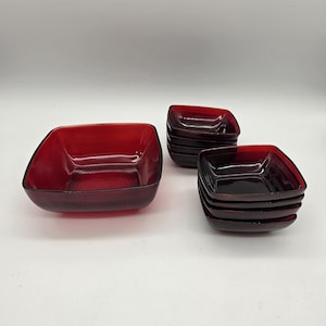 May include: A set of square red glass bowls consisting of one large bowl and two stacks of smaller bowls. Each bowl features a smooth, translucent red finish with a subtle, radiating pattern visible on the interior base of the larger bowl.