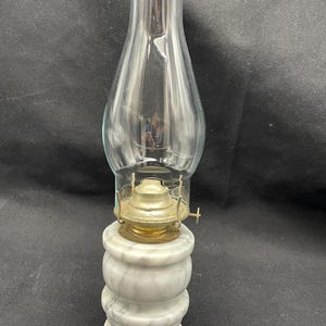 May include: An antique oil lamp with a clear glass chimney and a brass burner. The base is made of white marble with gray veining. The lamp is designed to sit on a flat surface. The lamp is a vintage item.