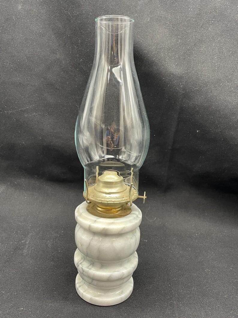 May include: An antique oil lamp with a clear glass chimney and a brass burner. The base is made of white marble with gray veining. The lamp is designed to sit on a flat surface. The lamp is a vintage item.