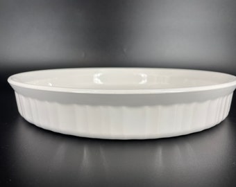 May include: A round white ceramic baking dish with a smooth interior and a fluted exterior design. The dish features a wide, flat rim and vertical ridges along the sides, providing a clean and functional look for oven-to-table serving and food preparation.