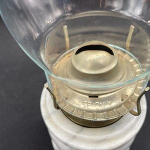 May include: Close-up of an antique oil lamp. The lamp features a clear glass globe, a brass burner assembly, and a white marble base. The burner is marked "USE ONLY".