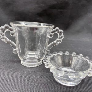 May include: A set of two clear glass cups with beaded edges. The larger cup has two handles and a rounded base. The smaller cup is a bowl shape.