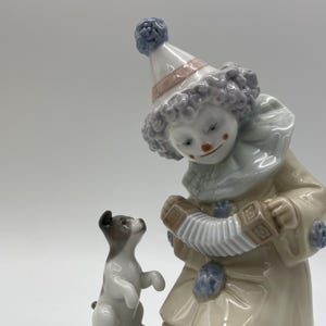 May include: A porcelain figurine of a clown wearing a blue and white hat playing an accordion. A small white dog with black spots stands on its hind legs next to the clown.