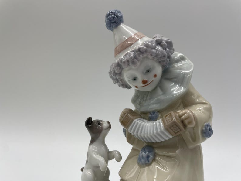 May include: A porcelain figurine of a clown wearing a blue and white hat playing an accordion. A small white dog with black spots stands on its hind legs next to the clown.
