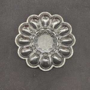 May include: Clear glass egg serving dish with a floral design etched in the center. The dish has a scalloped edge and individual egg-shaped compartments. A decorative piece for serving eggs.