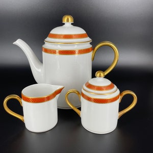 May include: A white porcelain tea set with gold accents and a red and gold stripe design. The set includes a teapot, creamer, and sugar bowl.