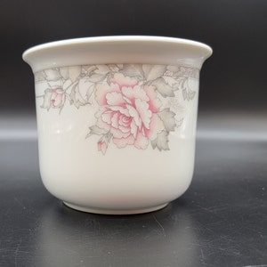 May include: White ceramic planter with a floral design featuring pink and gray roses. The planter has a rounded shape and a smooth finish.