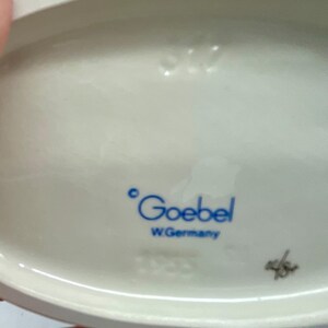 May include: Close-up of the base of a white porcelain figurine. The figurine is marked with the words "Goebel" and "W. Germany".