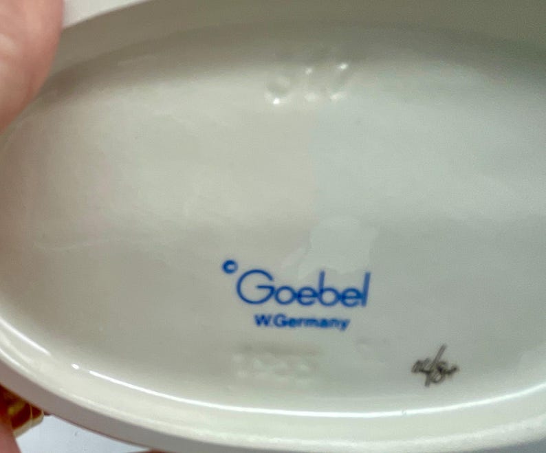 May include: Close-up of the base of a white porcelain figurine. The figurine is marked with the words "Goebel" and "W. Germany".
