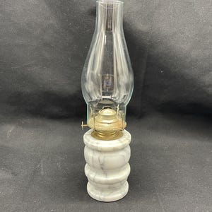 May include: A small oil lamp with a clear glass chimney and a marble base. The base is white with gray veining and has a stacked, rounded design. The lamp's metal components are gold-colored.