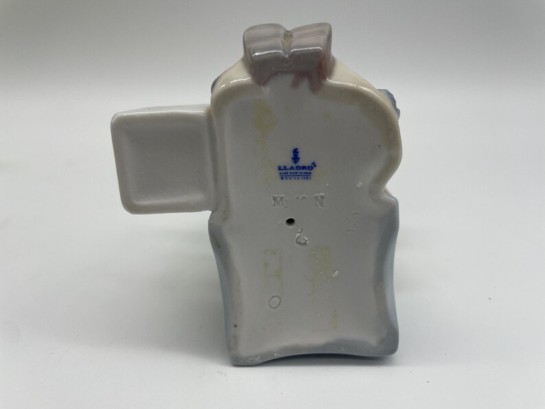 May include: A white porcelain figurine of a woman wearing a blue dress and a white hat. The figurine is holding a bouquet of flowers. The figurine is marked with the Lladro logo and the number 5710.