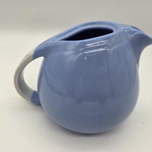 May include: A light periwinkle blue ceramic creamer or small pitcher with a rounded, bulbous body, a short pouring spout, and a curved handle. The exterior has a smooth, glossy finish, and the handle features a contrasting light gray accent.