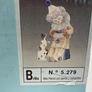 May include: A porcelain figurine of a clown holding an accordion, with a small dog sitting on a box beside it. The figurine is painted in shades of blue, white, and yellow. The box has the text "Brillo" and "N.&ordm; 5.279 Ni&ntilde;o Pierrot con perrito y concertina" printed on it.