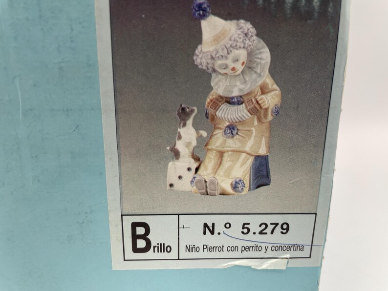 May include: A porcelain figurine of a clown holding an accordion, with a small dog sitting on a box beside it. The figurine is painted in shades of blue, white, and yellow. The box has the text "Brillo" and "N.&ordm; 5.279 Ni&ntilde;o Pierrot con perrito y concertina" printed on it.