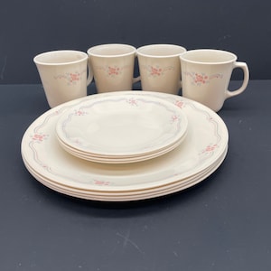 May include: A cream-colored dinnerware set featuring floral and ribbon designs in pink and blue. The set includes four mugs, four plates, and four smaller plates. The plates have a scalloped edge and a gold trim.