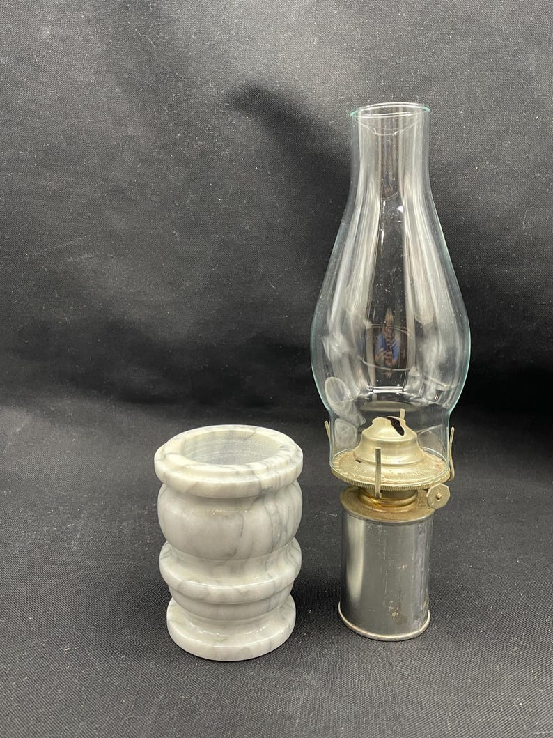May include: An antique oil lamp with a clear glass chimney and a metal base, next to a small, cylindrical marble container with a gray and white pattern. The lamp is unlit.