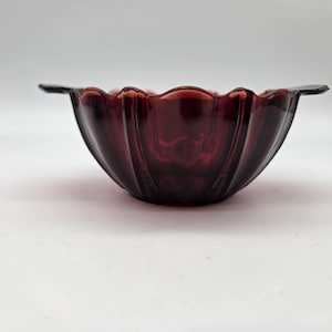 May include: A deep red glass bowl with a scalloped rim and two small handles. The bowl has vertical ridges and a glossy finish. The bowl is likely vintage and suitable for serving or display.