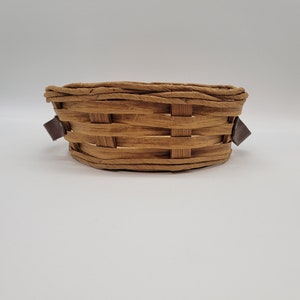 May include: A woven basket with a brown finish and a rectangular shape. The basket has a woven design with a braided rim and brown leather accents on each side.