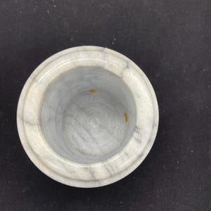 May include: A round, marble container with a light gray and white color scheme. The container has a circular opening and a slightly raised rim. The interior of the container is a lighter shade of gray with subtle veining.