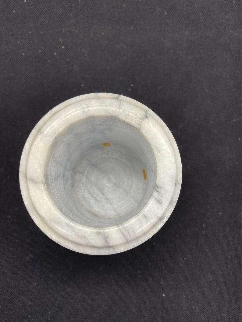 May include: A round, marble container with a light gray and white color scheme. The container has a circular opening and a slightly raised rim. The interior of the container is a lighter shade of gray with subtle veining.