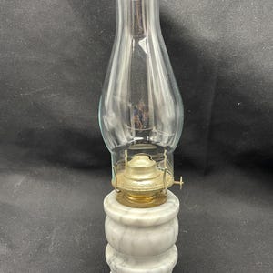 May include: A vintage oil lamp with a clear glass chimney and a brass burner. The lamp sits on a white marble base with a stacked, rounded design. The lamp is designed to hold oil for illumination.