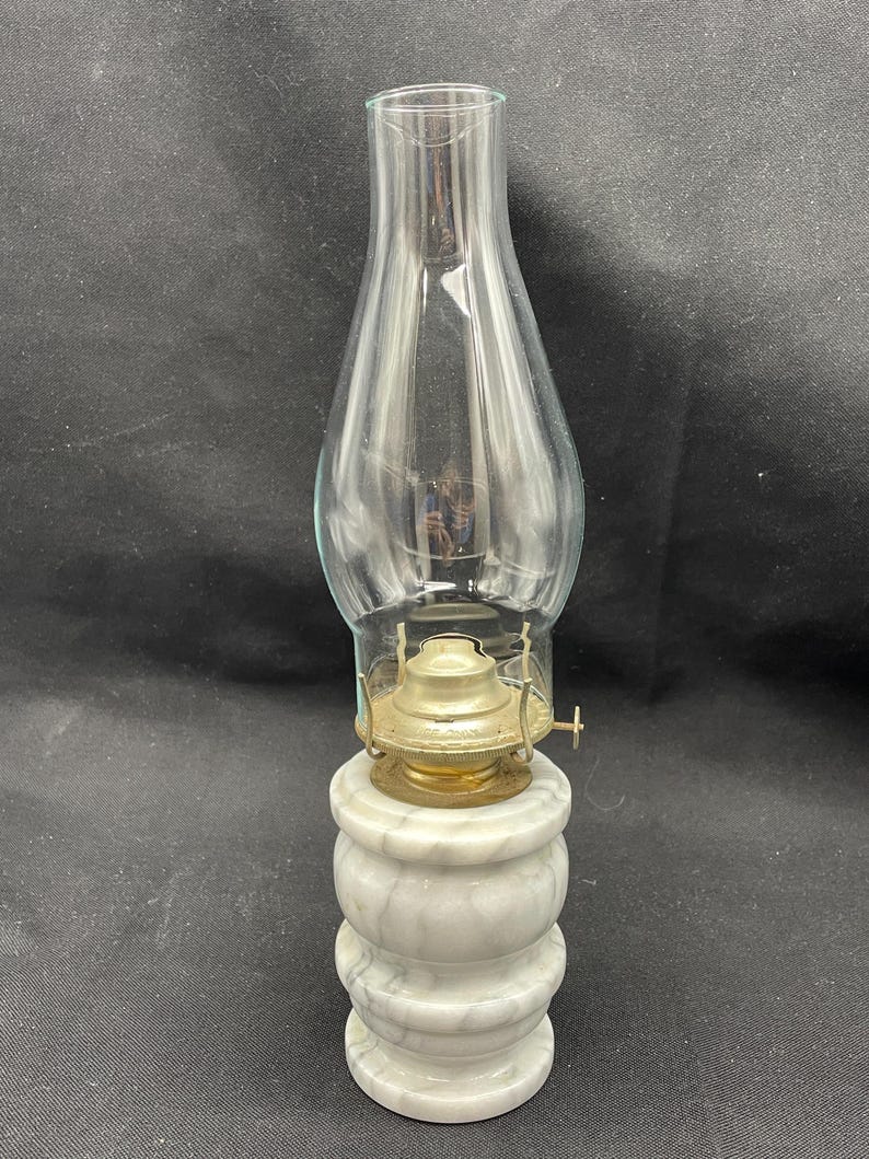 May include: A vintage oil lamp with a clear glass chimney and a brass burner. The lamp sits on a white marble base with a stacked, rounded design. The lamp is designed to hold oil for illumination.