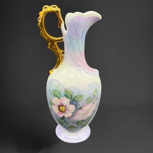 May include: A porcelain pitcher featuring a glossy white finish with iridescent pastel pink and blue highlights. The body is decorated with hand-painted pink flowers and green leaves. It includes a prominent, ornate, metallic gold handle.