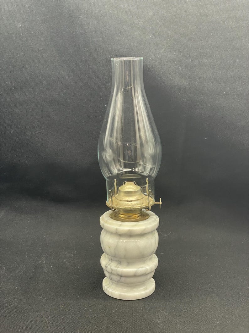 May include: A vintage oil lamp with a clear glass chimney and a marble base. The base is white with gray veining, and the lamp's metal components are gold-colored. The lamp is designed to sit on a flat surface.