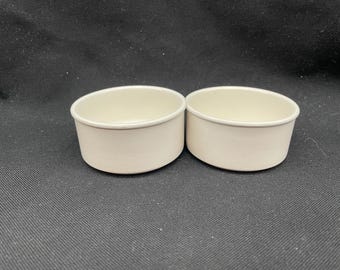 May include: Two off-white, oval-shaped ramekins. The small, ceramic bowls have a smooth, matte finish and a slightly rounded rim. They are placed side-by-side on a dark surface, ideal for baking or serving individual portions.