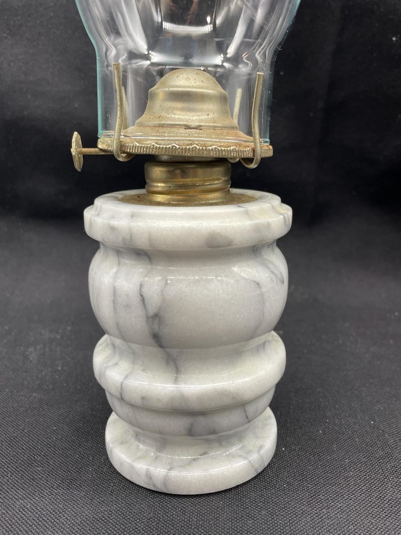 May include: A vintage oil lamp with a clear glass chimney and a marble base. The base is white with gray veining, and the metal components are brass and silver-toned. The lamp has a classic, antique aesthetic.