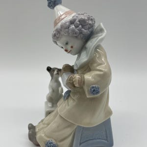 May include: A porcelain figurine of a clown with purple hair, wearing a blue and white costume, sitting on a blue stool and playing an accordion. A small white dog sits beside the clown.
