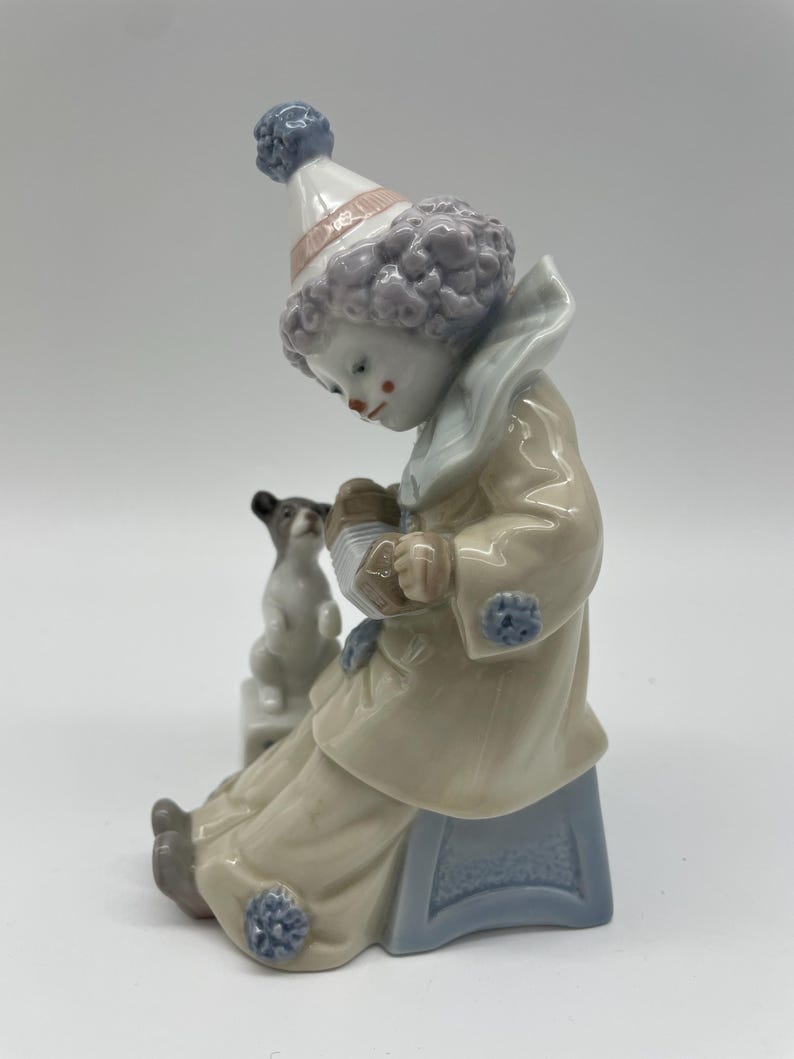 May include: A porcelain figurine of a clown with purple hair, wearing a blue and white costume, sitting on a blue stool and playing an accordion. A small white dog sits beside the clown.