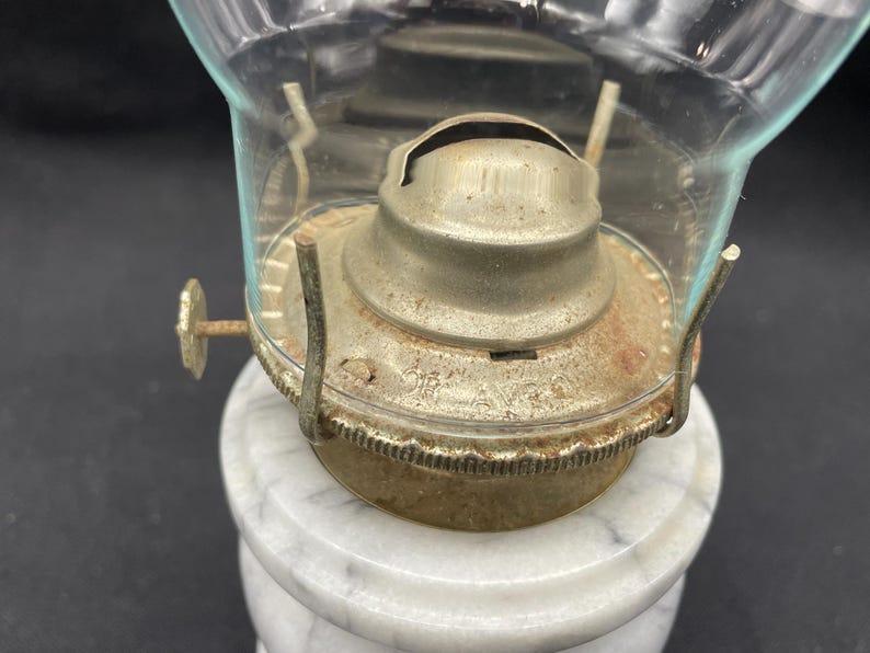 May include: Close-up of an antique oil lamp. The lamp features a clear glass globe and a metal burner assembly with visible rust. The base is made of white marble. The lamp is designed to sit on a table or other flat surface.