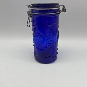 May include: A cobalt blue glass jar with a hinged lid and metal clasp. The jar features embossed floral and fruit designs. The jar is cylindrical and stands upright against a white background. The lid is dark blue.
