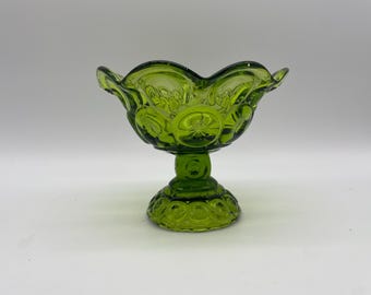 May include: A green glass pedestal bowl with a scalloped rim. The bowl has a star and moon design and sits on a round base. The bowl is likely used for serving or display.