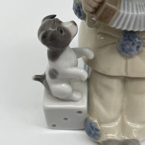 May include: A porcelain figurine of a boy wearing a blue and white outfit playing an accordion. A small dog sits on a white cube with black dots next to the boy.