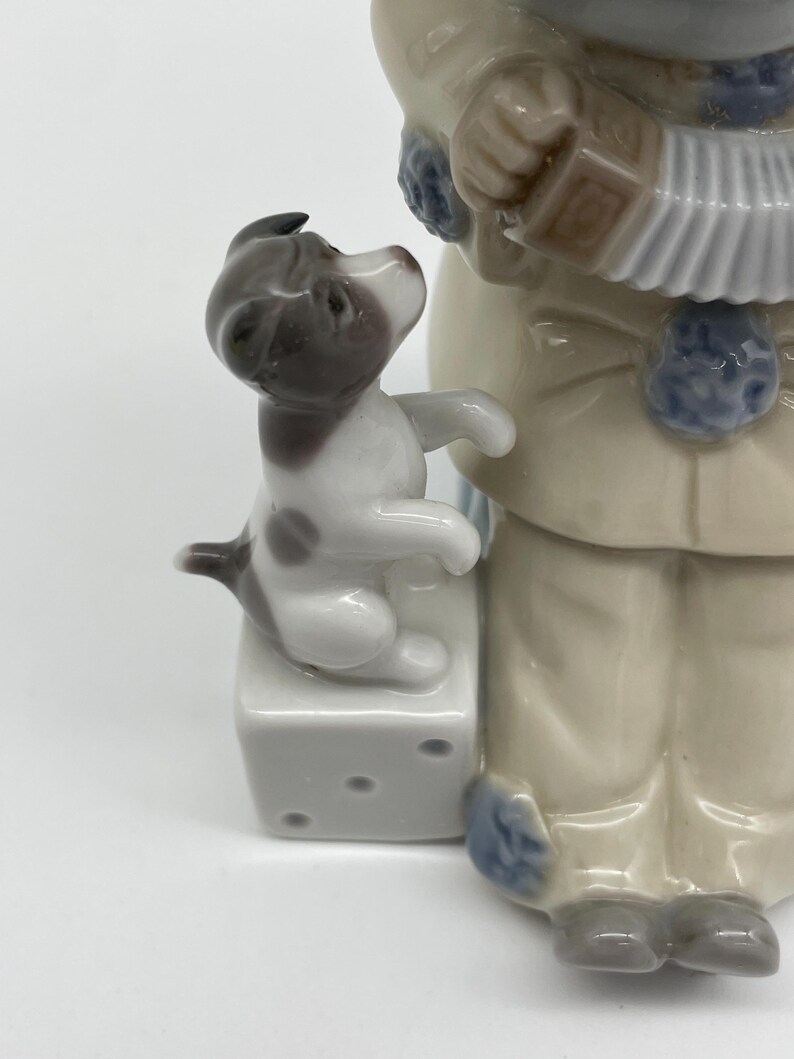 May include: A porcelain figurine of a boy wearing a blue and white outfit playing an accordion. A small dog sits on a white cube with black dots next to the boy.