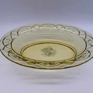 May include: A vintage, oval-shaped, amber-colored glass serving dish. The rim features a geometric design with floral accents. The center of the dish has a rose motif. The dish is likely from the Depression era.
