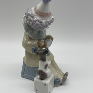 May include: A porcelain figurine of a clown with a blue and white hat, playing an accordion, and sitting on a blue and white bench with a small white and brown dog sitting on a white cube.