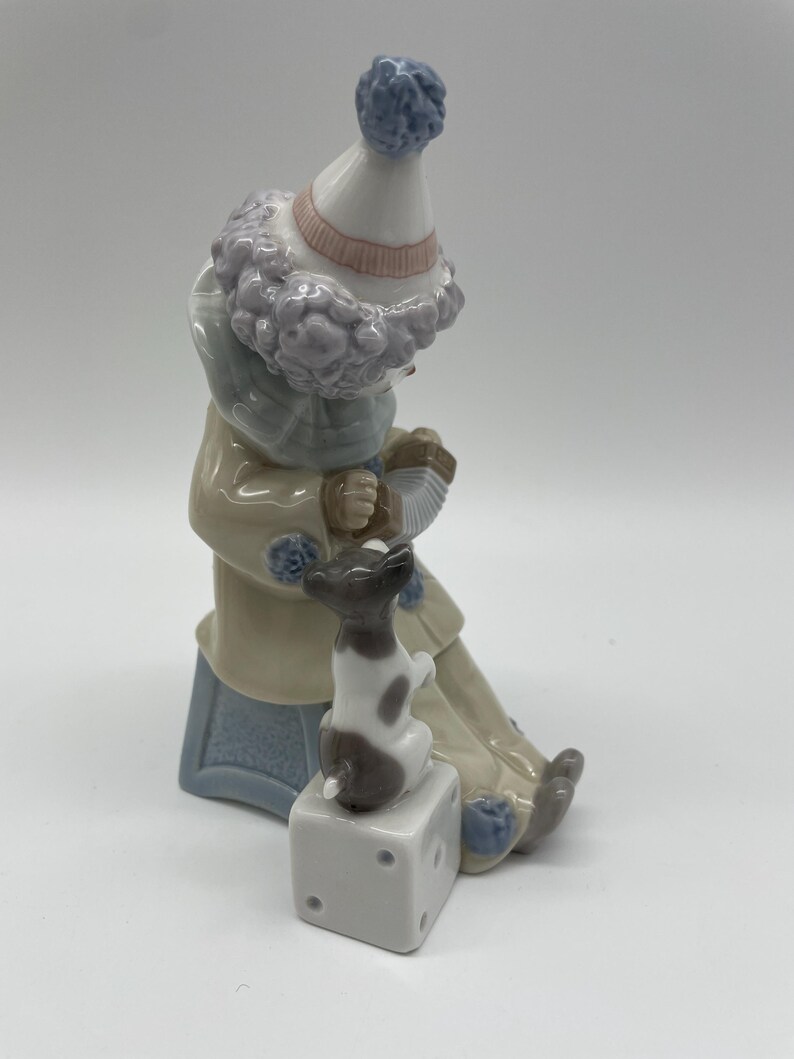 May include: A porcelain figurine of a clown with a blue and white hat, playing an accordion, and sitting on a blue and white bench with a small white and brown dog sitting on a white cube.